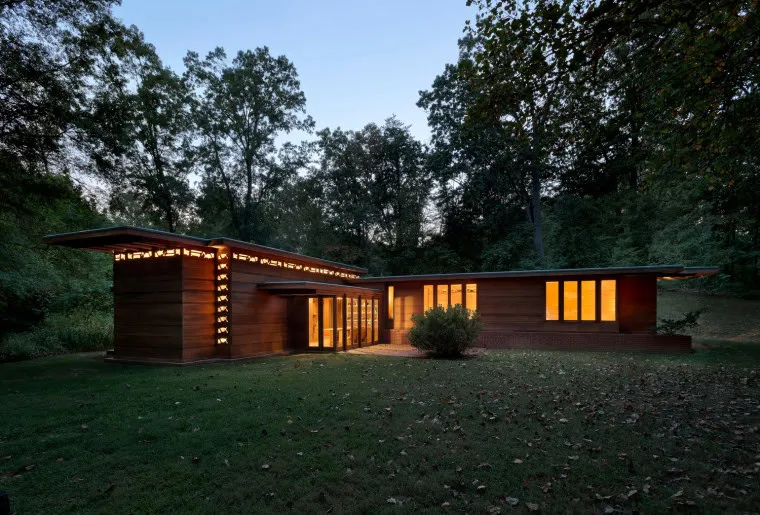 Modern, single-story house with large windows glowing with warm light at dusk, surrounded by trees and a grassy lawn, showcasing clean lines and natural wood exterior.