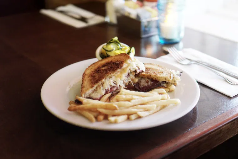 A plate with a grilled sandwich filled with meat and cheese, served with French fries and a small side of pickles, on a wooden table next to cutlery and condiments.