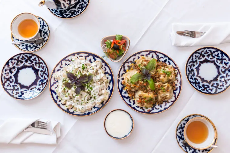 Overhead view of a table set with two ornate plates of food, a small bowl of sauce, a dish of salad, teacups with tea, and folded napkins on a white tablecloth.