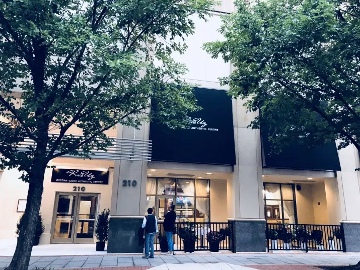 Two people stand outside the entrance of a building with green awnings labeled Rudy Boston Oral & Maxillofacial Surgery. The building is at 210 and is surrounded by trees and a sidewalk.