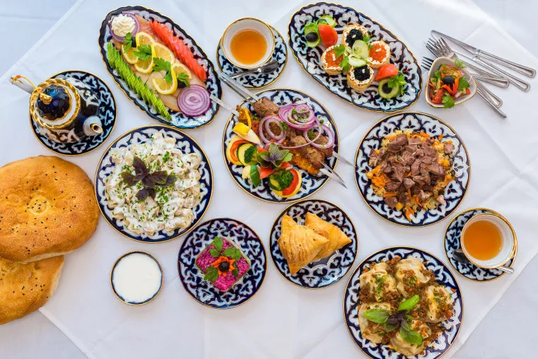 A table set with assorted dishes, including fresh salads, grilled meats, bread, colorful appetizers, tea in patterned cups, and sauces, all served on ornate plates over a white tablecloth.