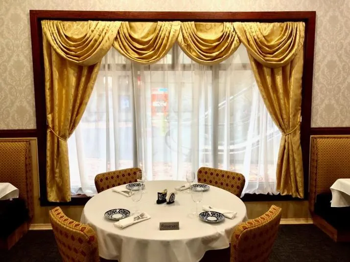 A small round dining table set for four sits by a window with gold drapes and white sheer curtains. The table has blue and white plates, glassware, and a Reserved sign. Upholstered chairs and booths surround the table.