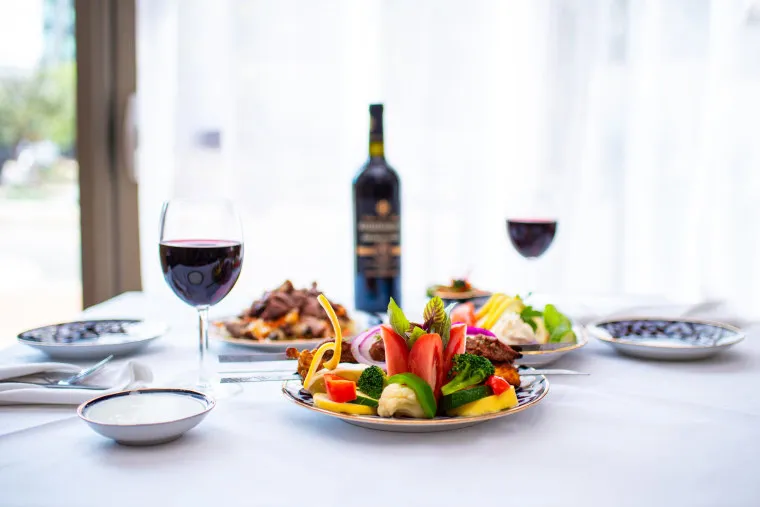 A table set with plates of colorful food, two glasses of red wine, and a bottle of wine in the center. The setting is bright with natural light coming through sheer curtains in the background.