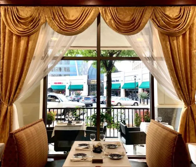 A restaurant dining area with elegant gold curtains and white sheer drapes framing a large window, through which a city street, parked cars, and storefronts are visible. Tables are set with plates, glasses, and cutlery.