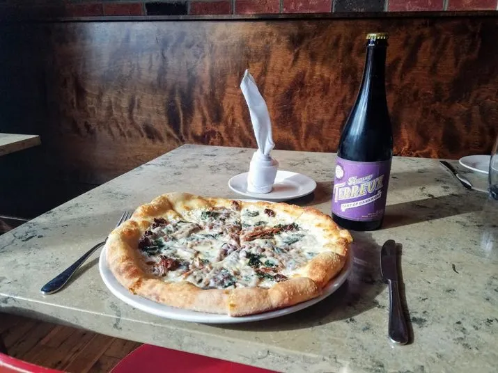 A pizza topped with greens and cheese sits on a plate with a fork and knife on either side. Nearby are a bottle of beer with a purple label and a plate with a white napkin standing upright.