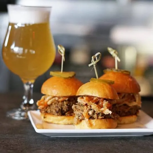 A glass of pale beer sits next to a square white plate holding three pulled pork sliders topped with pickles, served on brioche buns and secured with toothpicks.