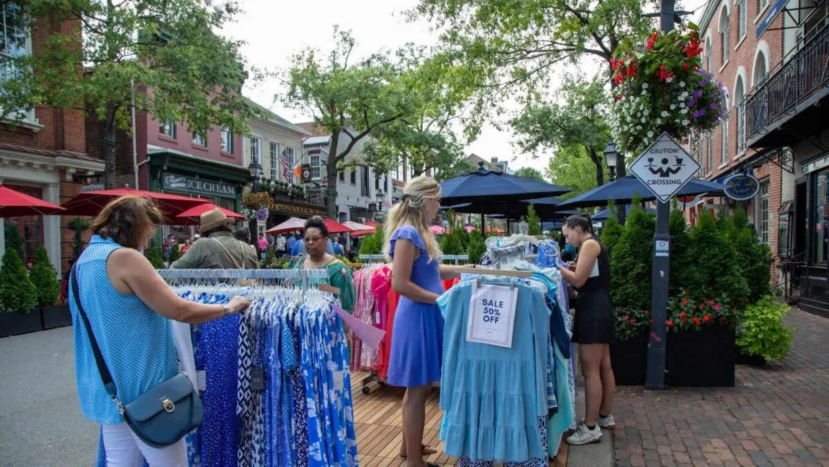 Women shop for dresses at outdoor clothing racks on a brick-lined street, with sale signs visible. Trees, shops, and people are in the background, creating a lively market atmosphere.