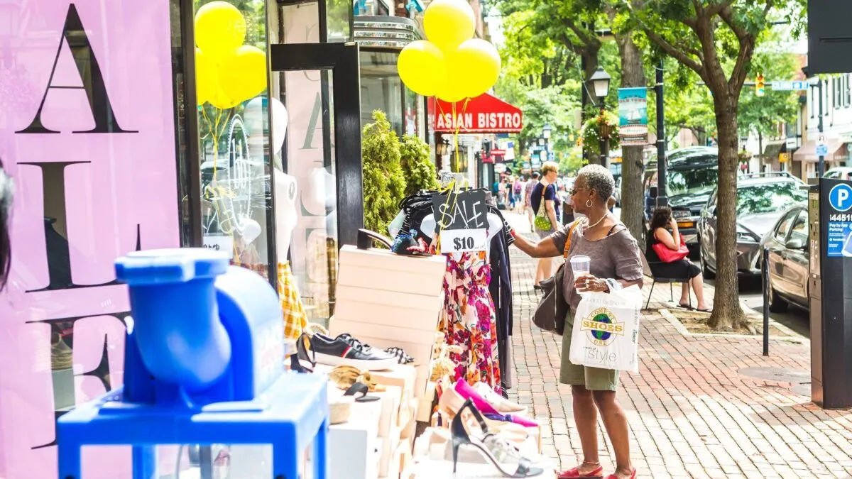 An older woman shops at an outdoor sidewalk sale, holding a shopping bag and looking at clothes on a rack marked “SALE $10.” Shoes and accessories are displayed nearby, with balloons and stores visible in the background.