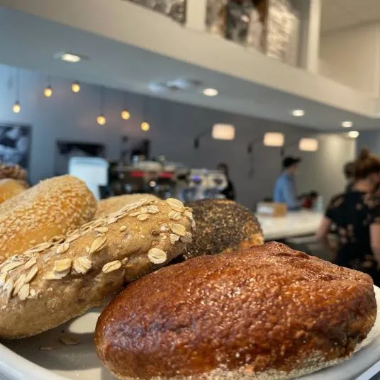 A close-up of assorted bread rolls, including seeded and oat-topped varieties, on a plate in a bright, modern café with people and hanging lights in the blurred background.