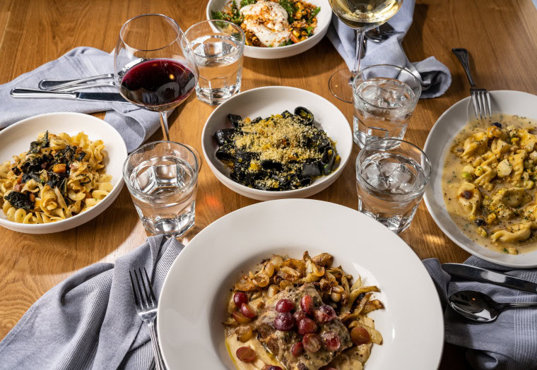 A wooden table set with plates of gourmet pasta, including squid ink pasta, creamy pasta, and fusilli, along with glasses of red and white wine, water, and a salad, with gray napkins and utensils.