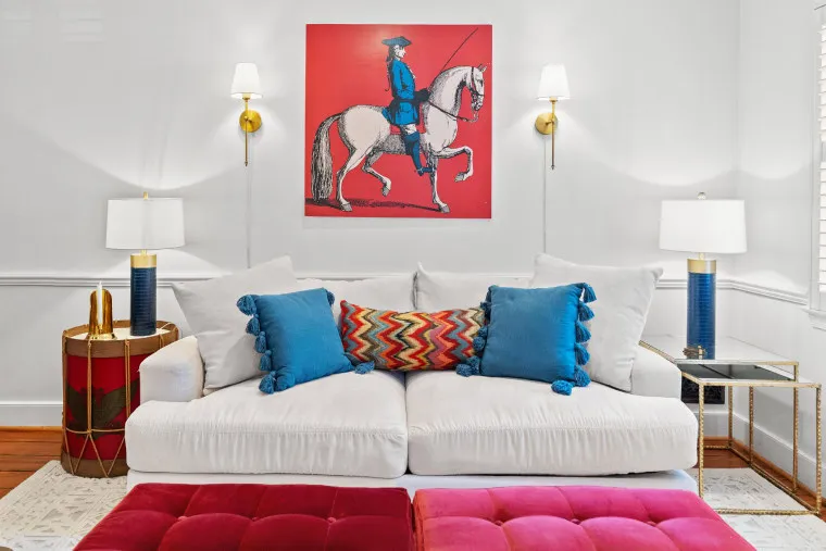 A modern living room with a white sofa, blue and red accent pillows, gold side tables, and wall lamps. A bold artwork of a rider on a horse with a red background hangs above the sofa.