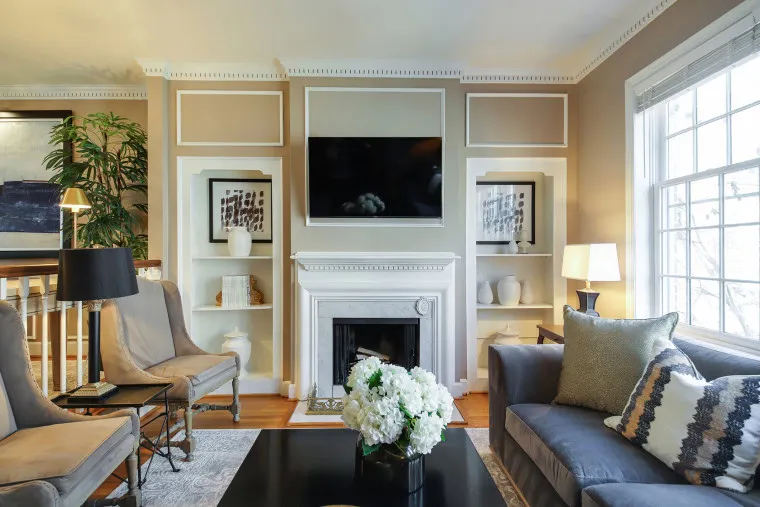 Bright living room with a fireplace, TV above the mantel, built-in shelves with decor, armchairs, a gray sofa, coffee table with white flowers, lamps, and large window letting in natural light.