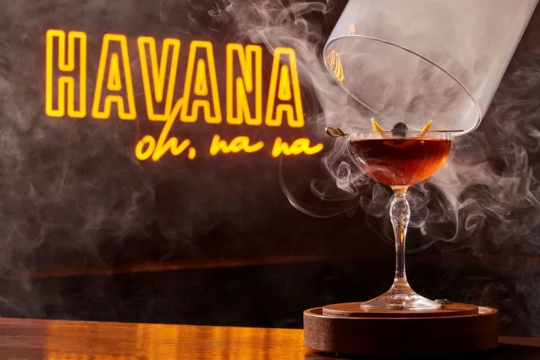 A smoky cocktail in a coupe glass sits on a wooden table, partially covered by a glass dome. In the background, neon signs read “HAVANA oh, na na” in yellow and orange lettering.
