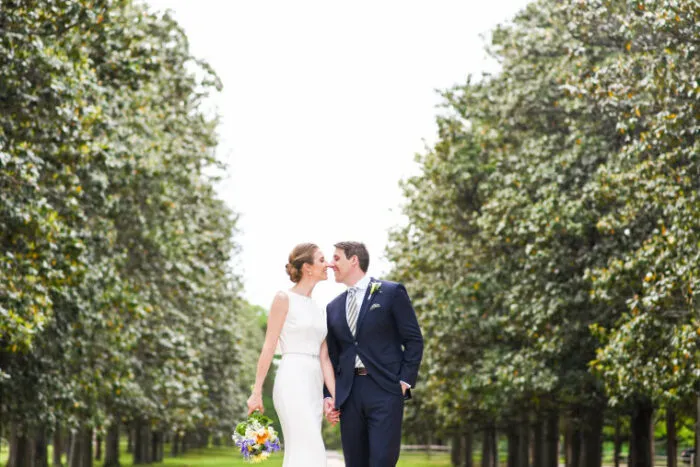 A bride and groom kiss while holding hands, standing on a path lined with tall, leafy trees. The bride wears a white dress and holds a bouquet; the groom wears a dark suit and tie.