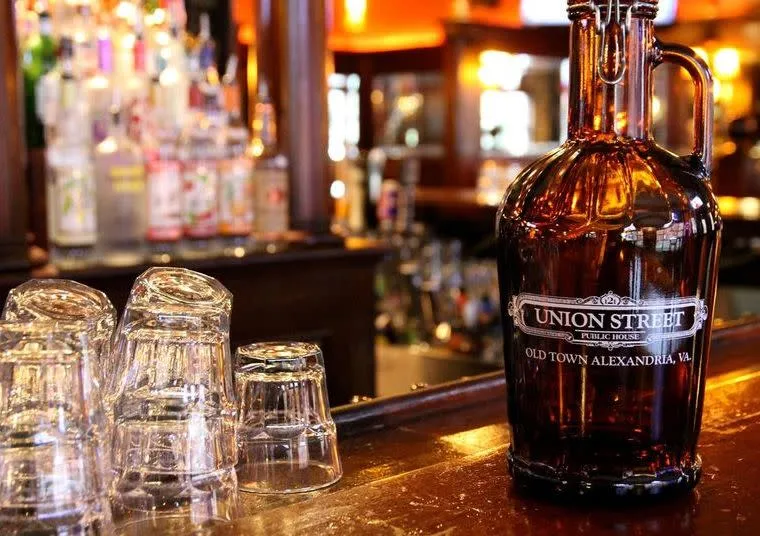 A dark glass growler labeled Union Street Public House, Old Town Alexandria, VA sits on a wooden bar beside several upside-down glass tumblers, with liquor bottles blurred in the background.