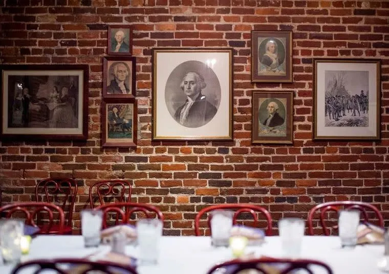 A brick wall decorated with framed portraits and historical images, mostly of men from the 18th century. In the foreground, a dining table is set with glasses, napkins, and red chairs.
