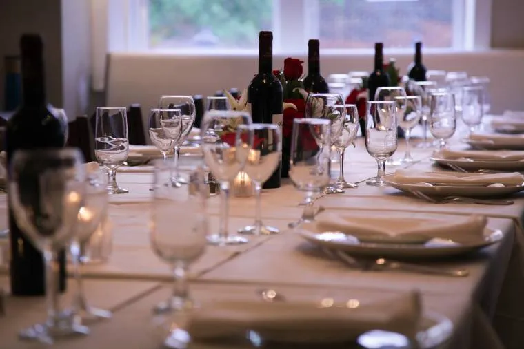 A formal dining table set with white tablecloths, neatly arranged wine glasses, plates, and silverware. Several bottles of red wine and decorative flowers are placed along the center of the table.