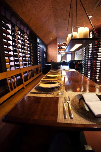 A long wooden dining table set with plates, cutlery, and glasses in a cozy restaurant, surrounded by shelves filled with bottles of wine and lit by soft candlelight.