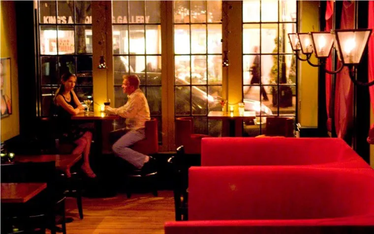 A dimly lit café with red booths and two people sitting at a small table near large windows, talking. Streetlights and people are visible outside through the windows at night.