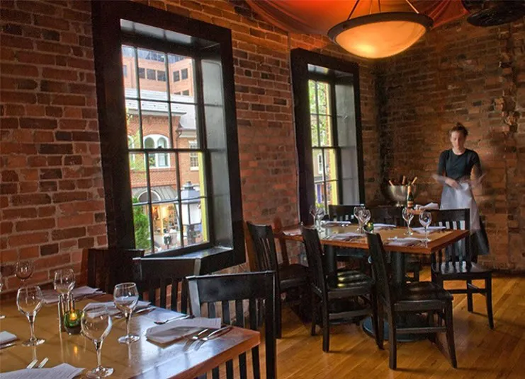 A cozy restaurant with brick walls, large windows, wooden tables set with glasses and napkins, and a server standing near a table. Natural light streams in from the windows, showing a city view outside.