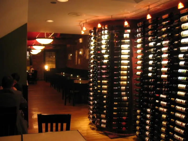 Dimly lit wine bar interior with tall racks filled with wine bottles along one wall, dark wooden tables and chairs, and soft, warm lighting creating a cozy atmosphere.