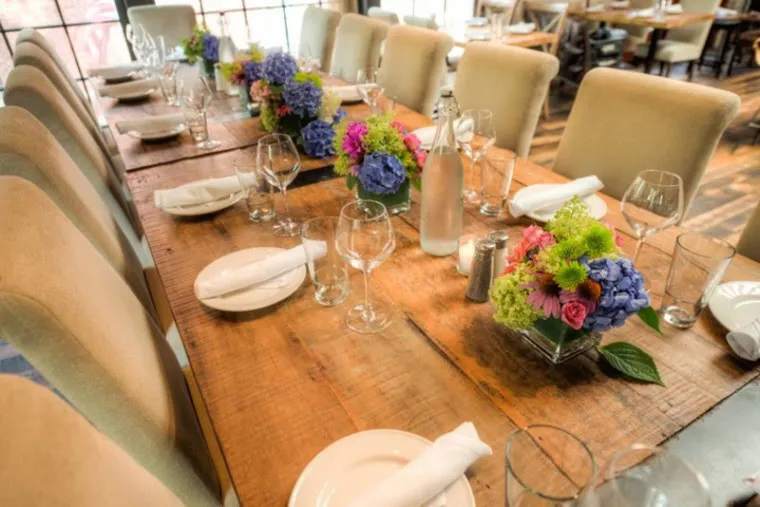 A long wooden dining table set for a meal, with beige chairs, white plates, glasses, napkins, and centerpieces of vibrant hydrangeas and other flowers in square vases. Sunlight streams in from large windows nearby.
