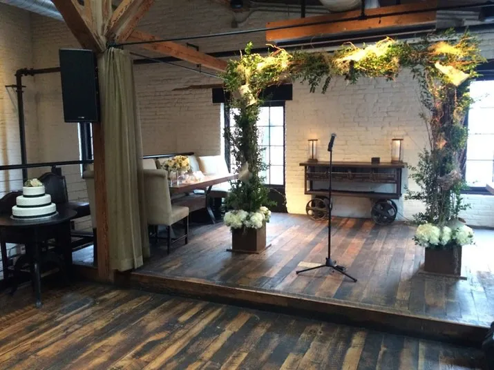 A wooden wedding arch decorated with greenery and flowers stands on a small indoor stage with a microphone. A tiered wedding cake is on a table to the left, and a console table with candles is at the back.