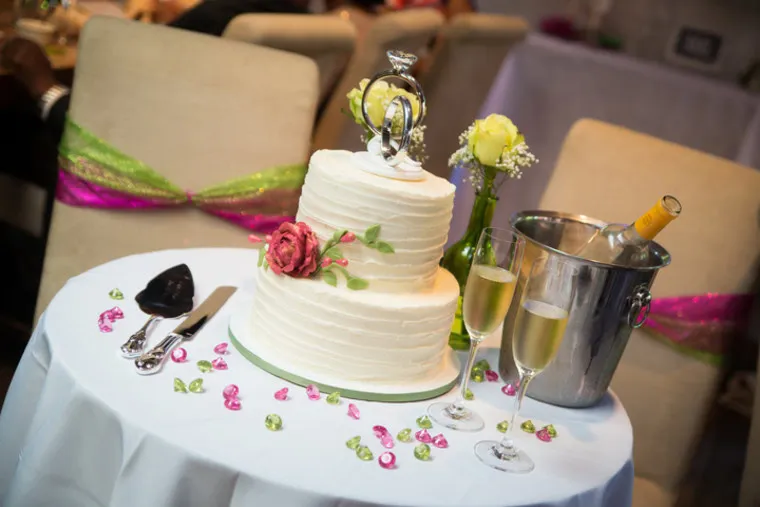 A white, two-tiered wedding cake with a pink flower and decorative topper sits on a table with two filled champagne glasses, a champagne bottle in an ice bucket, and scattered pink and green decorations.