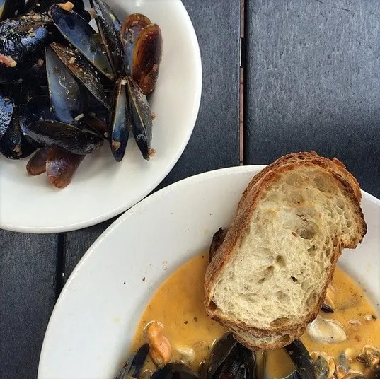 A plate of mussels in orange sauce with a slice of crusty bread sits next to a dish of empty mussel shells on a dark wooden table.