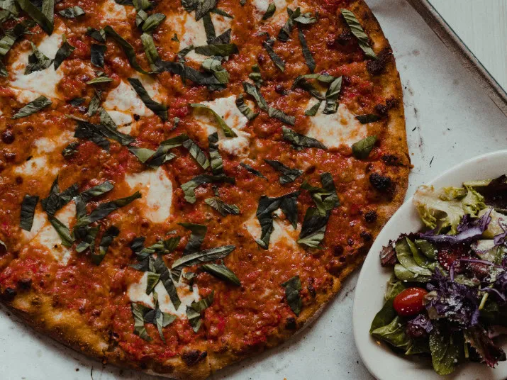 A close-up of a margherita pizza topped with tomato sauce, mozzarella cheese, and fresh basil, served on a tray next to a small plate of mixed greens salad with cherry tomatoes.