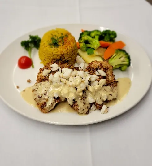 A white plate with breaded fish topped with creamy sauce and crab meat, served with yellow rice, steamed broccoli, carrots, zucchini, a cherry tomato, and a parsley garnish.