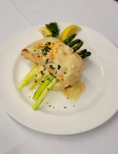 A plated dish featuring a piece of baked fish topped with creamy sauce and herbs, served over mashed potatoes and asparagus spears, garnished with parsley and lemon wedges.