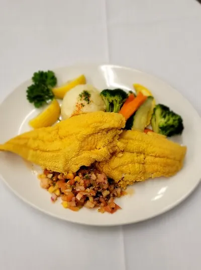A plate with two pieces of fried fish, a scoop of mashed potatoes, steamed vegetables, lemon wedges, parsley garnish, and a small serving of diced salsa on a white tablecloth.