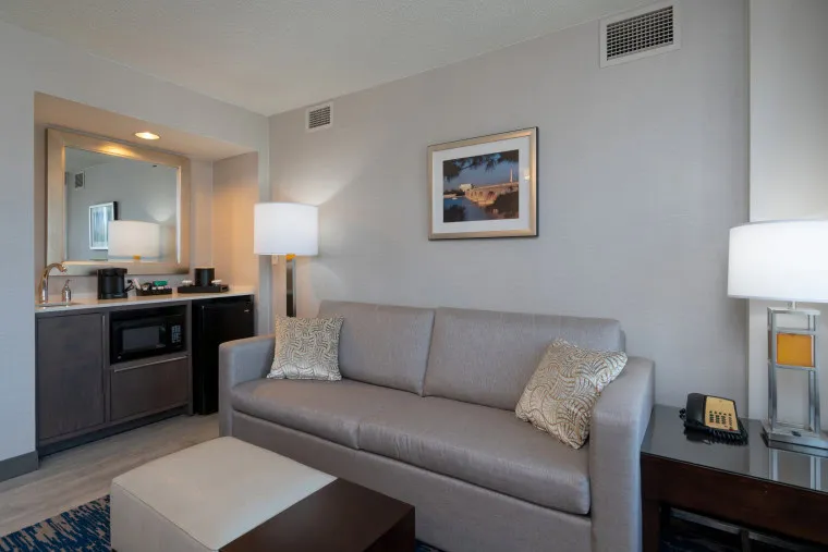 A modern hotel room features a gray sofa with two decorative pillows, a side table with a lamp and phone, a large ottoman, a kitchenette area with a microwave and mini fridge, and framed wall art above the sofa.