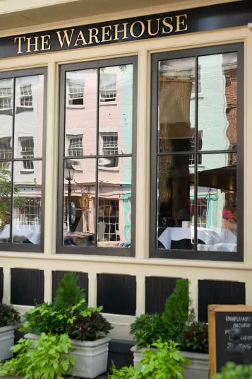 Exterior of The Warehouse restaurant, showing large windows with reflections of pastel-colored buildings across the street, some indoor tables visible, and potted plants along the sidewalk.