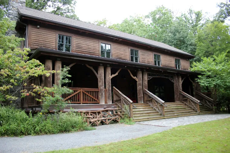A large rustic wooden building with a covered porch, wooden railings, and steps leading up to the entrance, surrounded by trees and greenery.