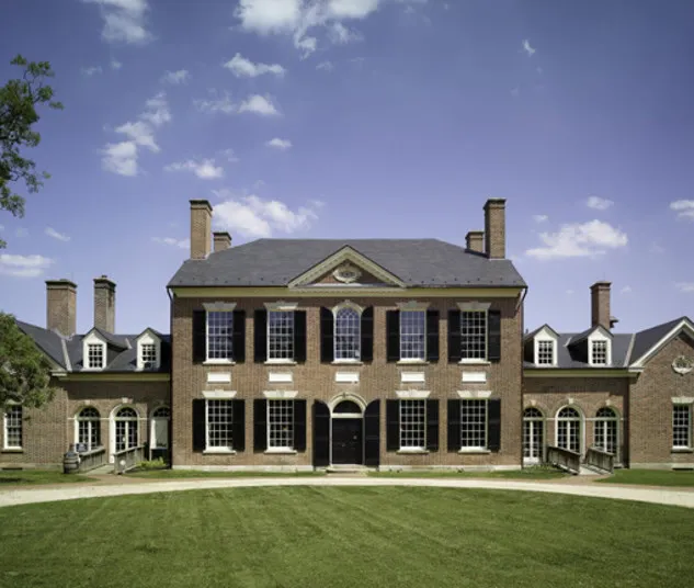 A large, symmetrical brick mansion with tall chimneys, black shutters, and white-trimmed windows stands under a blue sky. A wide green lawn leads up to the main entrance, which features double doors and arched windows.