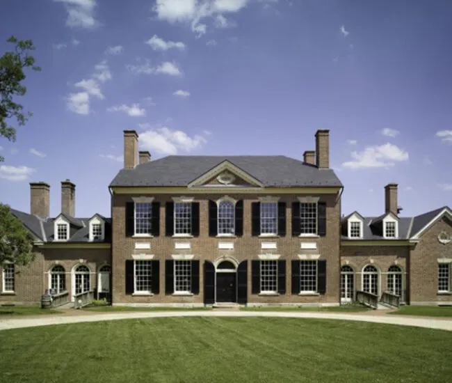 A large, symmetrical brick mansion with black shutters, multiple chimneys, and arched windows, sits under a blue sky with scattered clouds, facing a manicured lawn.