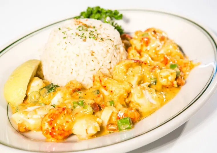 A plate with a serving of white rice garnished with herbs, a wedge of lemon, and a creamy seafood dish with shrimp, green onions, and sauce, garnished with parsley on a white plate.