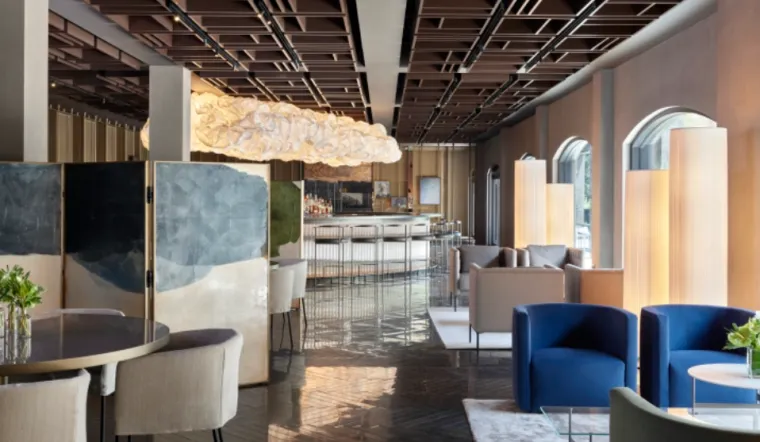 A modern lounge features stylish seating, a curved bar with stools, large arched windows, soft lighting, and artistic room dividers. A sculptural cloud-like light fixture hangs from the ceiling.