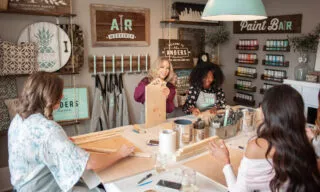 Four women sit around a table in a craft workshop, smiling and working on wooden projects. Paint supplies and brushes are on the table, with colorful paint jars and signs displayed on the walls behind them.