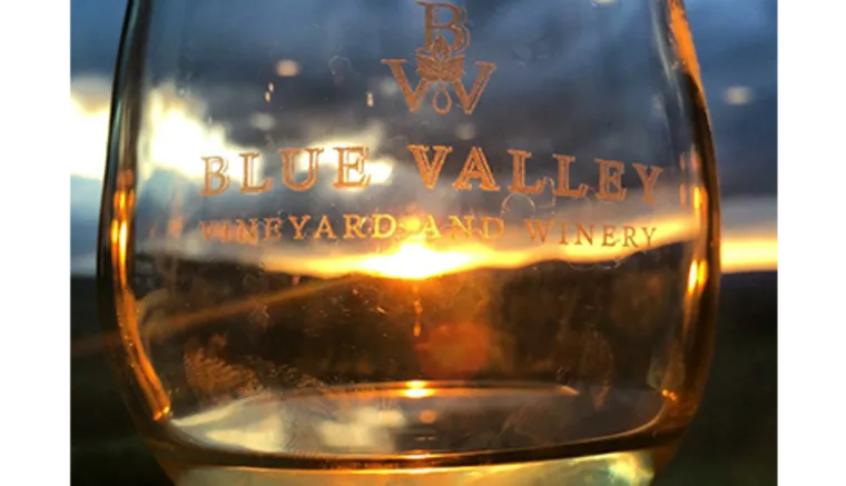 A close-up of a wine glass with Blue Valley Vineyard and Winery etched on it, held up against a sunset, with golden light shining through the glass.