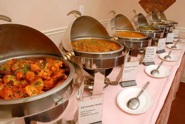 A buffet table with five metal chafing dishes holding various Indian dishes, including Tandoori Murg. Plates and serving spoons are placed in front of each dish on a pink tablecloth.