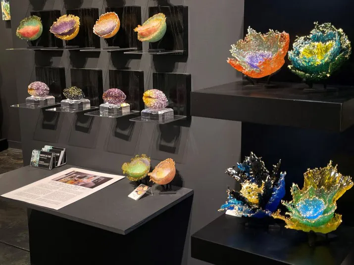 Colorful, textured glass sculptures resembling abstract flowers or leaves are displayed on black shelves and a table in a gallery setting, with exhibition labels and an artist statement visible nearby.