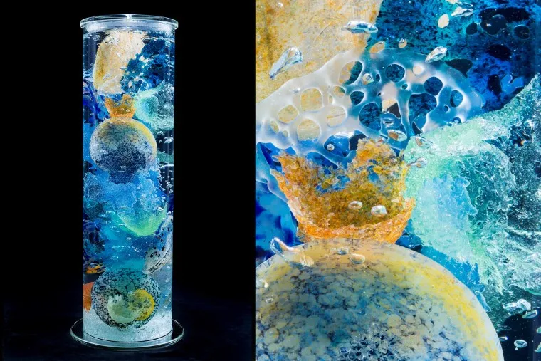 A tall, clear cylindrical glass sculpture filled with colorful abstract shapes in blue, yellow, and orange. Close-up shows textured glass bubbles and swirling patterns against a black background.