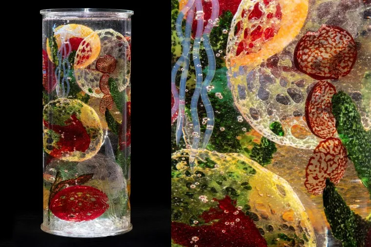A tall, clear cylindrical sculpture contains colorful, textured shapes resembling fruit and abstract forms, suspended in transparent material with bubbles. A close-up shows intricate details and vibrant colors inside the cylinder.