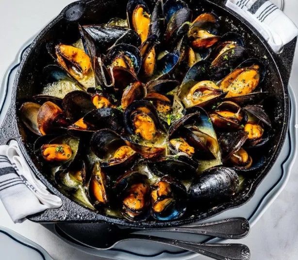 A cast iron skillet filled with cooked mussels in a creamy sauce, garnished with herbs. The skillet rests on a white plate with two cloth napkins and silverware beside it.