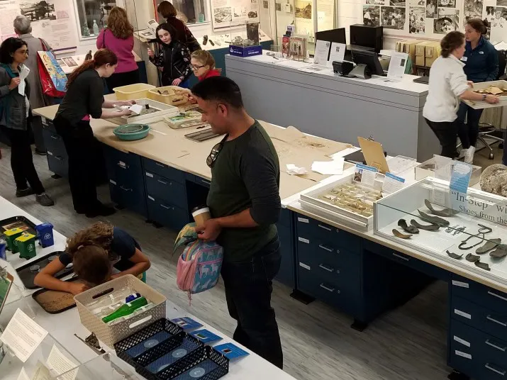 People explore and interact with artifacts and displays at a museum exhibit, with several adults and children working on activities at tables and examining objects in a busy, well-lit room.