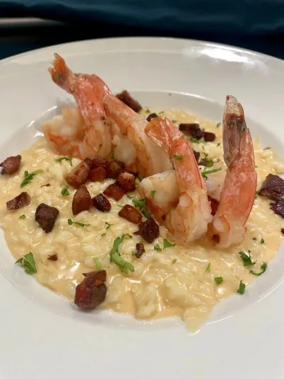 A creamy plate of shrimp and grits, topped with grilled shrimp, diced pieces of bacon, and fresh herbs, served in a white bowl.