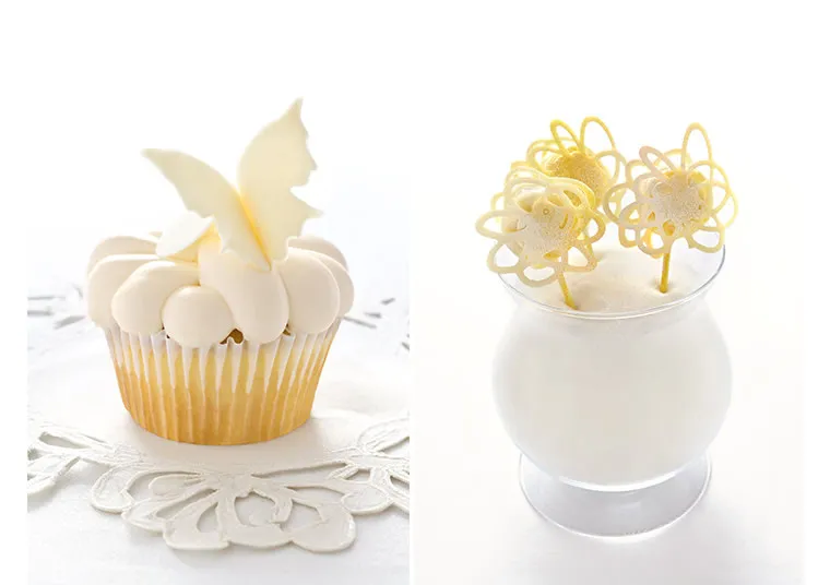 A vanilla cupcake with white frosting and a white chocolate butterfly, next to a glass containing three decorative yellow sugar lollipops shaped like flowers, both displayed on a white surface.